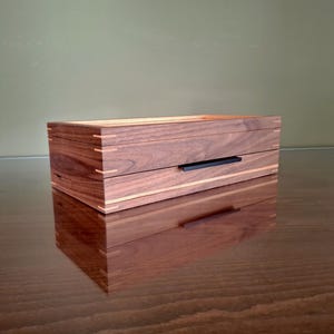May include: A rectangular wooden box featuring dark walnut wood grain, light-colored wood inlay accents, and visible corner joinery. The box includes a single drawer with a thin, horizontal black metal handle centered on the front panel.