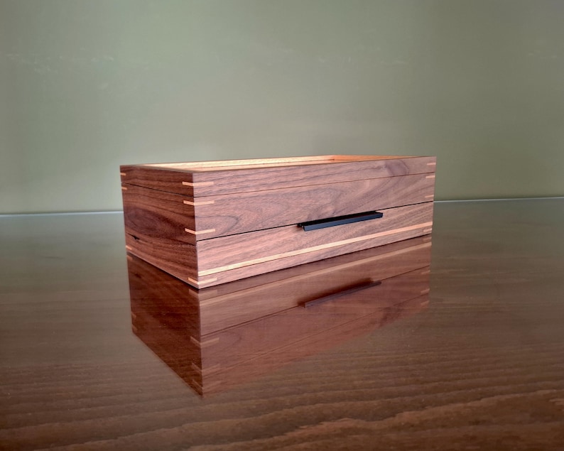May include: A rectangular wooden box featuring dark walnut wood grain, light-colored wood inlay accents, and visible corner joinery. The box includes a single drawer with a thin, horizontal black metal handle centered on the front panel.