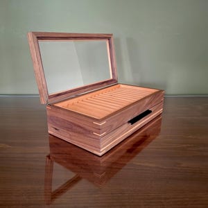 May include: A rectangular wooden display box with a hinged glass lid and a dark brown finish. The interior features multiple parallel slots for organizing items, and the exterior corners are reinforced with light-colored wood joinery details.