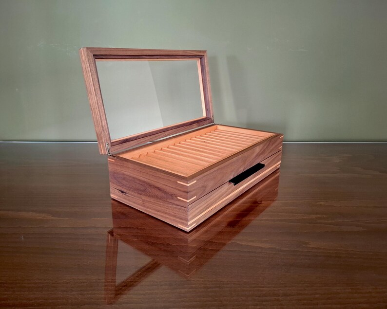 May include: A rectangular wooden display box with a hinged glass lid and a dark brown finish. The interior features multiple parallel slots for organizing items, and the exterior corners are reinforced with light-colored wood joinery details.