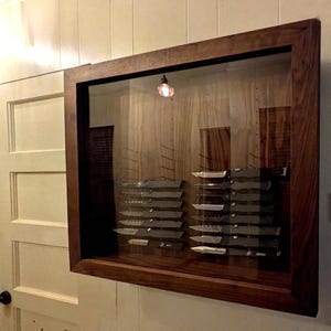 Op de afbeelding: Een rechthoekige houten vitrinekast met een glazen voorzijde, gemonteerd aan een muur. Binnenin zijn twee verticale kolommen van zakmessen gerangschikt op een houten achterpaneel. Het frame is gemaakt van donkerbruin hout met een zichtbare nerf.