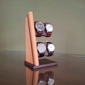 Op de afbeelding: Een houten horlogestandaard met vier polshorloges. De standaard is gemaakt van licht en donker hout, met twee niveaus om de horloges vast te houden. De horloges hebben leren bandjes en zilverkleurige wijzerplaten. De standaard staat op een reflecterend oppervlak.