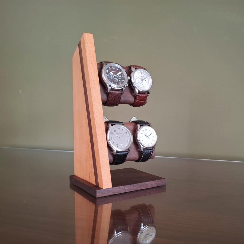 Op de afbeelding: Een houten horlogestandaard met vier polshorloges. De standaard is gemaakt van licht en donker hout, met twee niveaus om de horloges vast te houden. De horloges hebben leren bandjes en zilverkleurige wijzerplaten. De standaard staat op een reflecterend oppervlak.