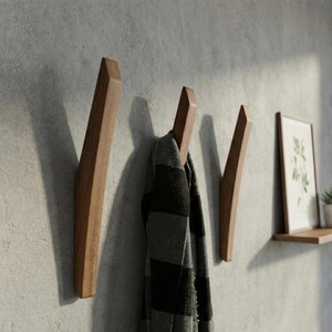 Mid-century modern solid wood wall hooks in a natural walnut finish, mounted on a textured light gray wall, with a black and gray plaid scarf hanging from the center hook, styled in a minimalist hallway with a floating wooden shelf and framed botanical print