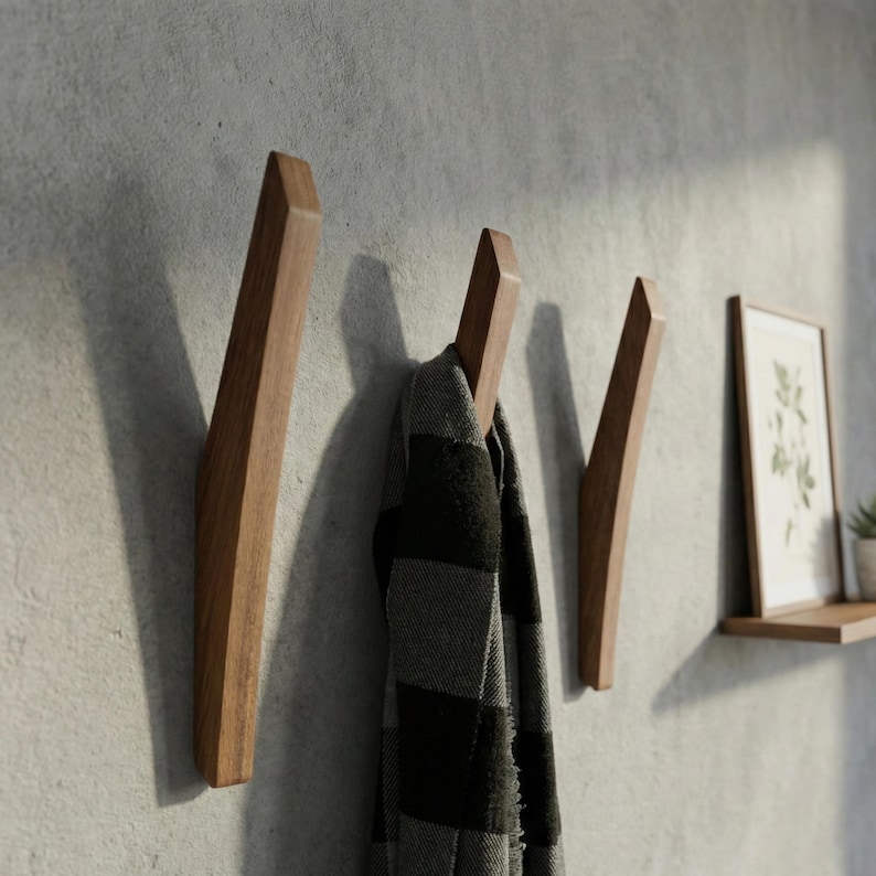 Mid-century modern solid wood wall hooks in a natural walnut finish, mounted on a textured light gray wall, with a black and gray plaid scarf hanging from the center hook, styled in a minimalist hallway with a floating wooden shelf and framed botanical print