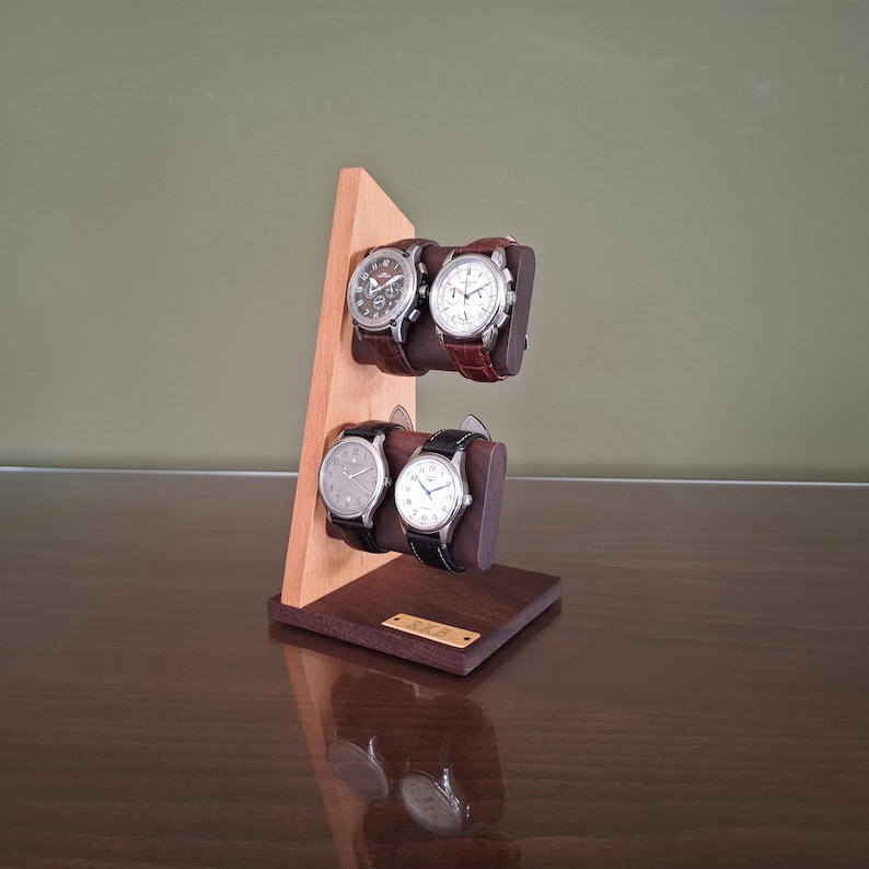 Op de afbeelding: Een houten horlogestandaard met vier polshorloges. De standaard is gemaakt van licht en donkerbruin hout, met twee niveaus om de horloges vast te houden. De horloges hebben leren bandjes en zilveren of zwarte wijzerplaten.