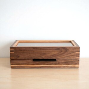 May include: A rectangular wooden storage box featuring a dark brown walnut grain, a clear glass top panel, and light-colored wood inlay accents. The box includes a single pull-out drawer with a thin, horizontal black metal handle centered on the front.