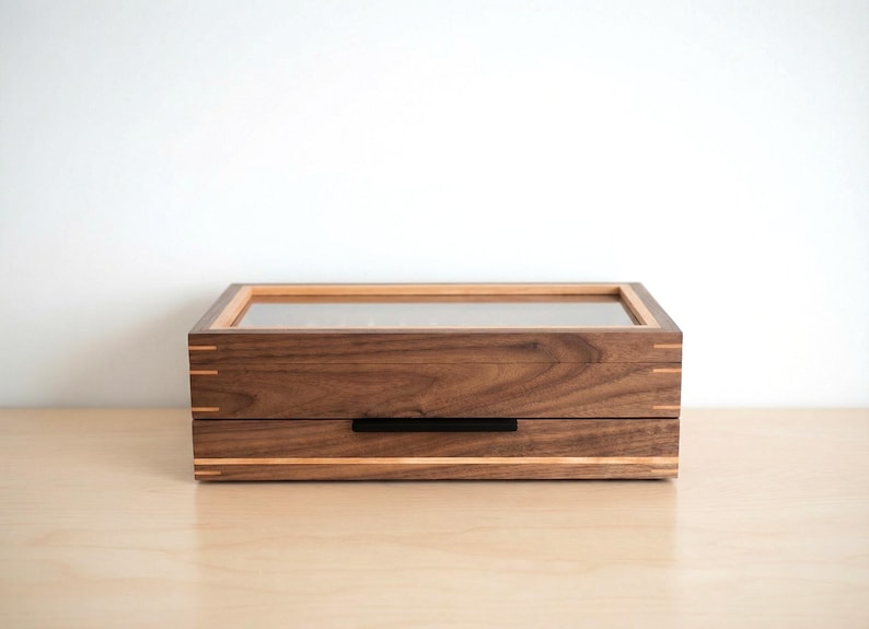 May include: A rectangular wooden storage box featuring a dark brown walnut grain, a clear glass top panel, and light-colored wood inlay accents. The box includes a single pull-out drawer with a thin, horizontal black metal handle centered on the front.