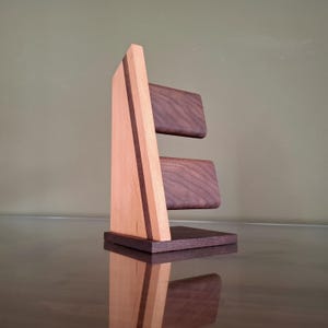 Op de afbeelding: Een houten horlogestandaard met een licht houten frame en donkere houten accenten. De standaard heeft twee donkere houten platforms voor het vasthouden van horloges. Het ontwerp omvat een donkere houten basis en een verticale lichte houten steun met een donkere houten streep.