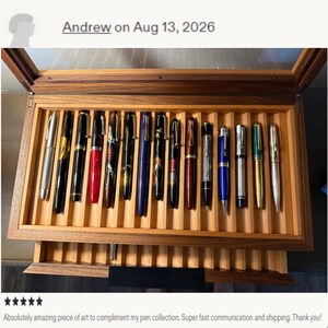 May include: A wooden display case with a glass lid containing fourteen fountain pens arranged in individual slots. The pens feature various colors and finishes, including silver, black, red, blue, and green, with gold or silver-toned clips.