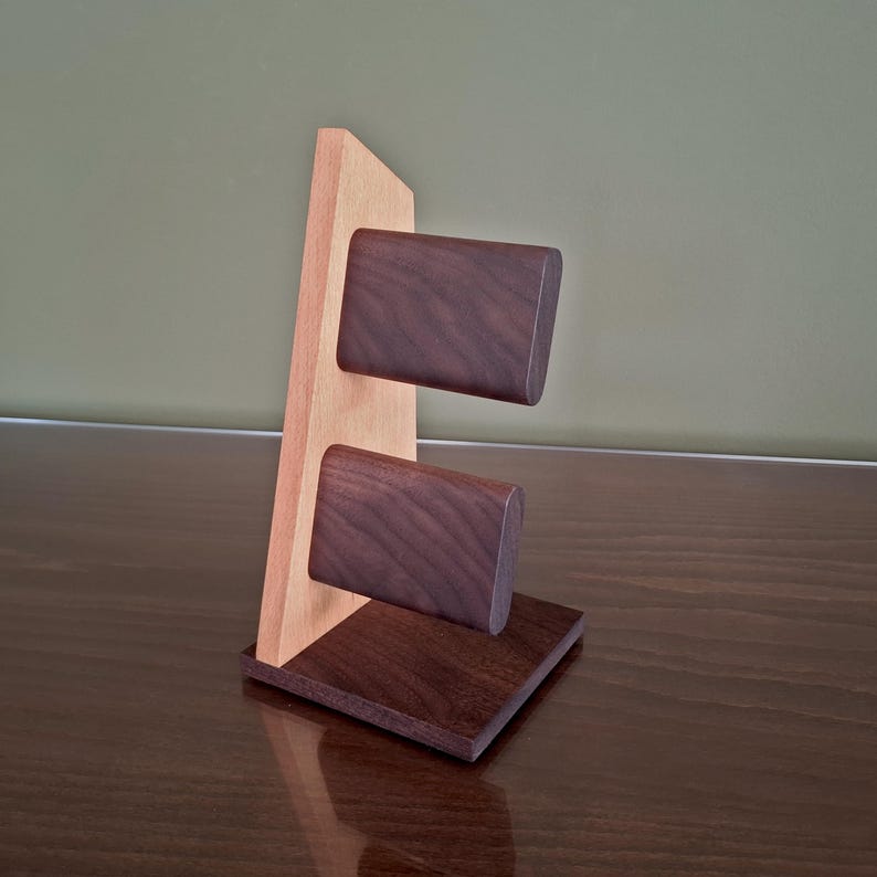 Op de afbeelding: Een houten displaystandaard met een lichtgekleurde verticale achterwand en twee donkerbruine rechthoekige blokken. De basis is een donkerbruin vierkant. De standaard is ontworpen om items vast te houden of weer te geven.