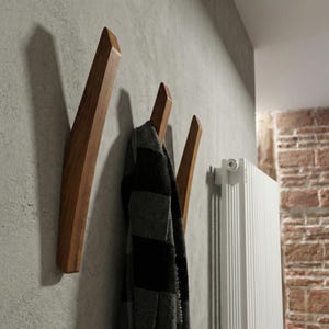 Mid-century modern solid wood wall hooks in a dark walnut finish, mounted on a textured concrete-style wall, with a black and gray scarf hanging from one hook, styled in a contemporary hallway interior with a white radiator and exposed brick detail.