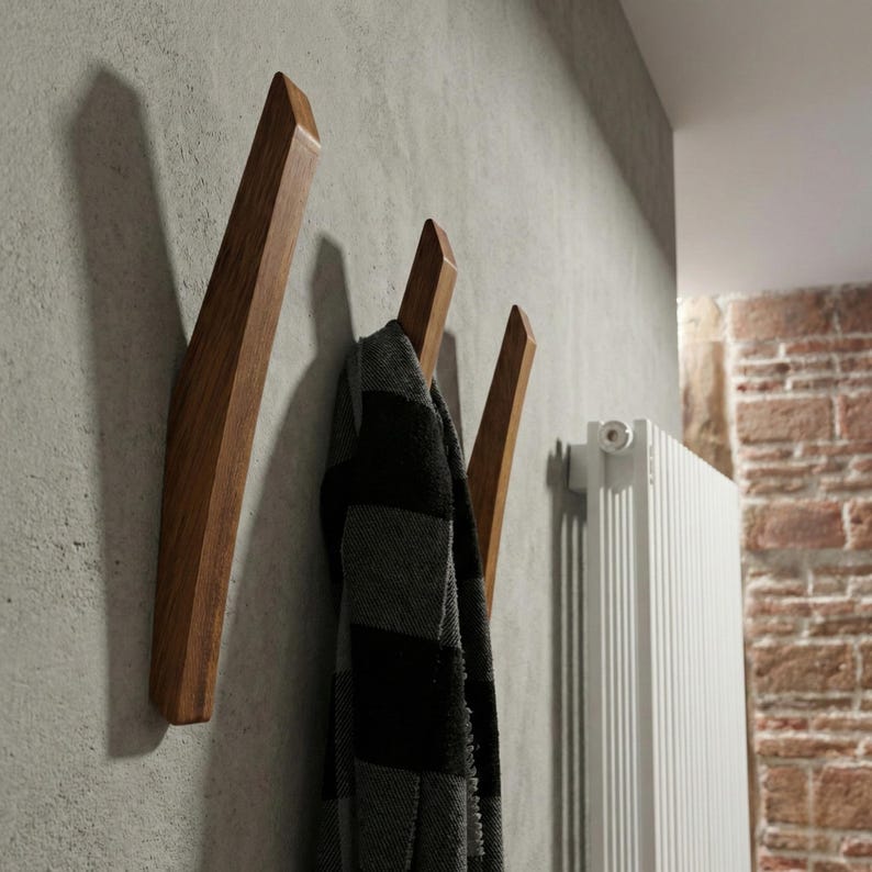 Mid-century modern solid wood wall hooks in a dark walnut finish, mounted on a textured concrete-style wall, with a black and gray scarf hanging from one hook, styled in a contemporary hallway interior with a white radiator and exposed brick detail.