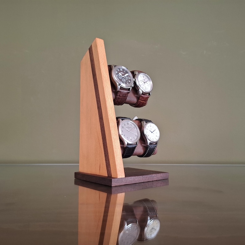 Op de afbeelding: Een houten horlogestandaard met een lichtbruine afwerking en een donkerbruine accentstreep. De standaard biedt plaats aan vier polshorloges met leren en metalen bandjes. De horloges hebben zilverkleurige wijzerplaten.