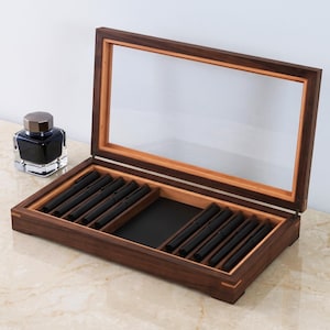 May include: A rectangular dark wood display case with a hinged glass lid, featuring interior slots for ten black pens and a central black rectangular compartment. Beside the case sits a square glass ink bottle with a dark metallic cap.