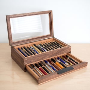 May include: A dark brown wooden display case with a hinged glass lid and a pull-out drawer. The interior features multiple grooved slots holding a collection of various fountain pens in assorted colors, patterns, and metallic finishes.