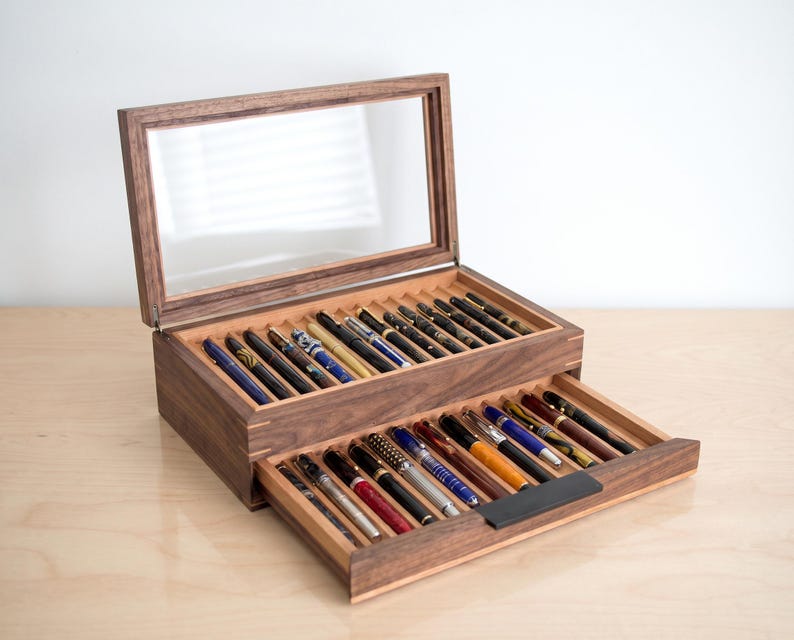 May include: A dark brown wooden display case with a hinged glass lid and a pull-out drawer. The interior features multiple grooved slots holding a collection of various fountain pens in assorted colors, patterns, and metallic finishes.