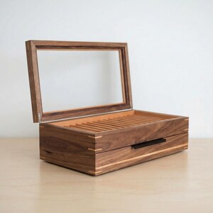 May include: A rectangular wooden display box crafted from dark brown walnut wood with a hinged lid featuring a clear glass window. The interior contains multiple parallel slots for organizing items, and the front includes a black horizontal handle.