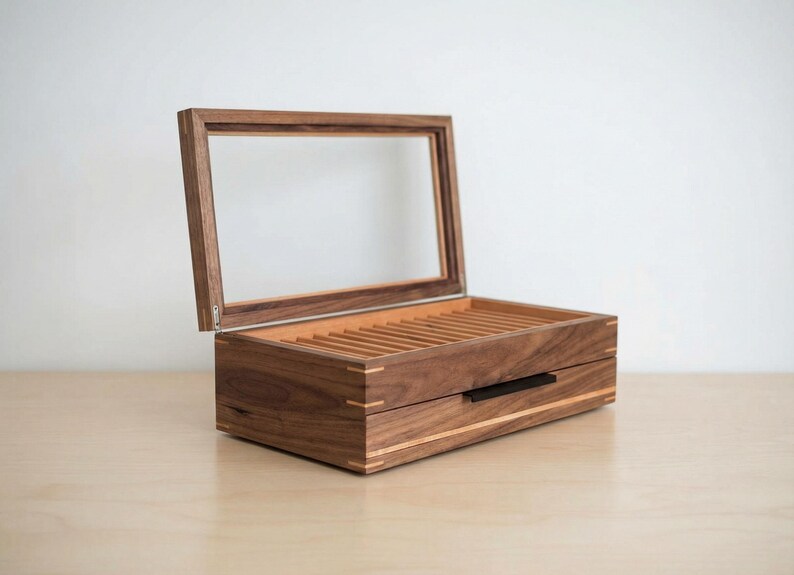 May include: A rectangular wooden display box crafted from dark brown walnut wood with a hinged lid featuring a clear glass window. The interior contains multiple parallel slots for organizing items, and the front includes a black horizontal handle.