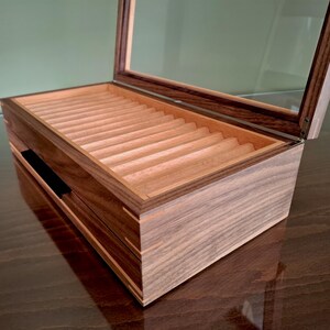May include: A rectangular wooden display box with a dark brown exterior and a light tan interior featuring multiple parallel grooves. The box has a hinged lid with a clear glass panel, allowing for a view of the interior storage compartments.