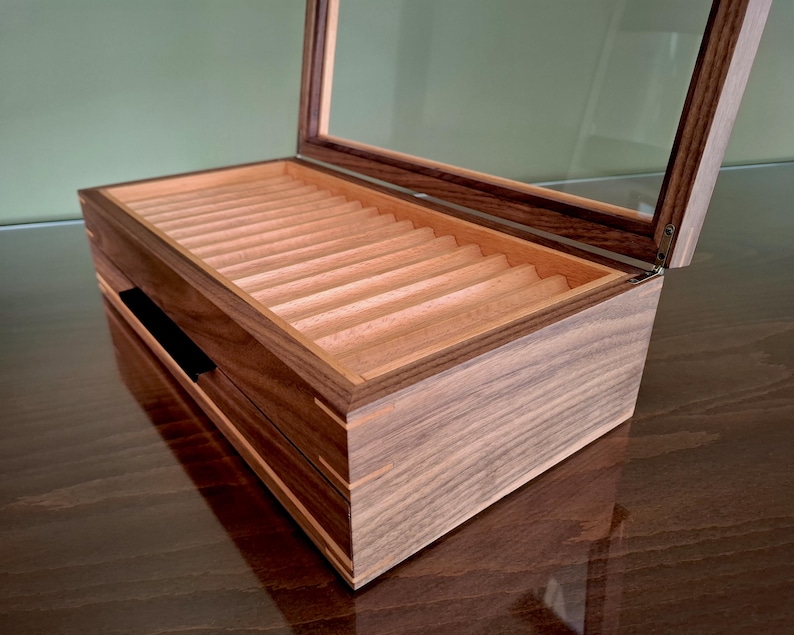 May include: A rectangular wooden display box with a dark brown exterior and a light tan interior featuring multiple parallel grooves. The box has a hinged lid with a clear glass panel, allowing for a view of the interior storage compartments.