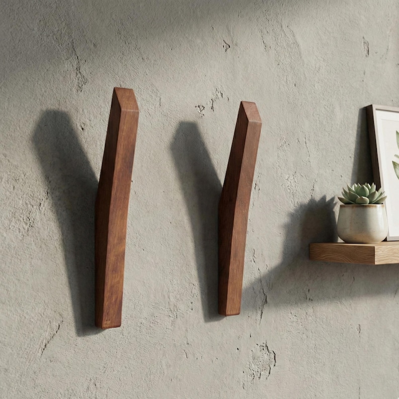 Mid-century modern solid wood wall hooks in a dark walnut finish, mounted on a textured light gray wall, featuring two sculpted angled wooden hooks with clean lines, styled with a floating wooden shelf, framed art, and a small potted plant.