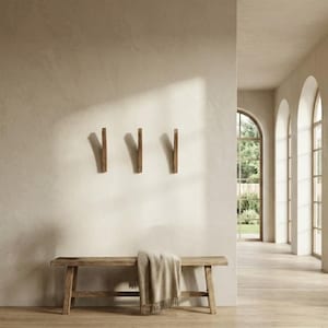 Minimalist entryway interior featuring three mid-century modern solid wood wall hooks in a natural oiled finish mounted on a warm neutral wall, styled above a rustic wooden bench with a draped throw blanket, in a bright space with arched windows and natural light.