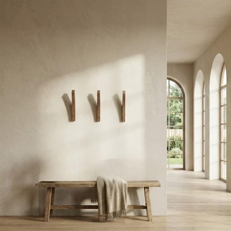 Minimalist entryway interior featuring three mid-century modern solid wood wall hooks in a natural oiled finish mounted on a warm neutral wall, styled above a rustic wooden bench with a draped throw blanket, in a bright space with arched windows and natural light.
