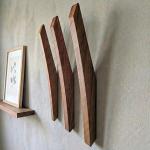 Angled mid-century modern wooden wall hooks in dark walnut finish mounted on a textured light gray wall, showing three sculpted solid wood hooks with visible grain, styled with a floating wooden shelf, framed botanical print