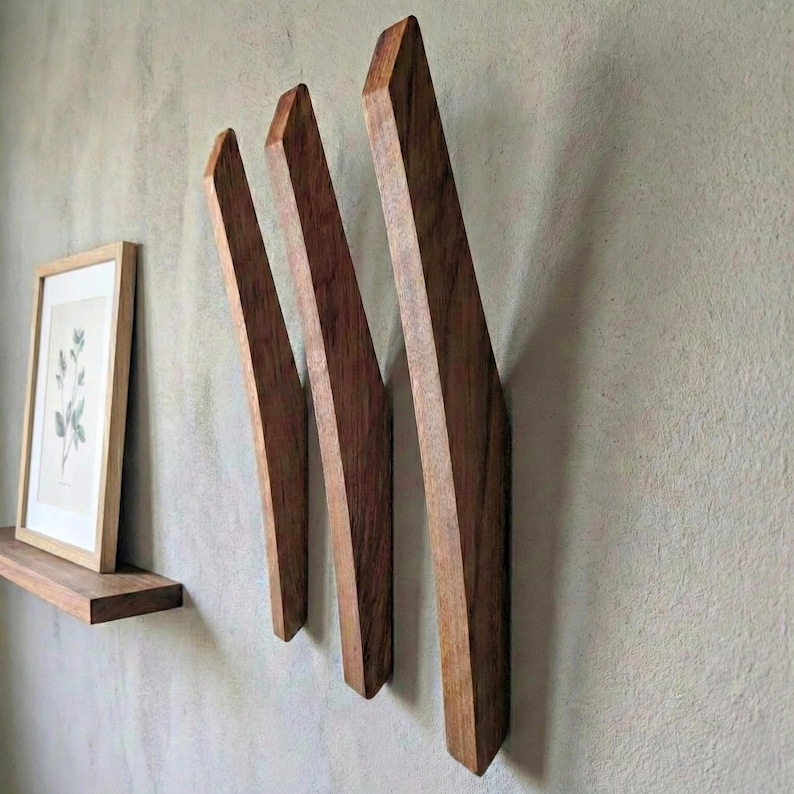 Angled mid-century modern wooden wall hooks in dark walnut finish mounted on a textured light gray wall, showing three sculpted solid wood hooks with visible grain, styled with a floating wooden shelf, framed botanical print