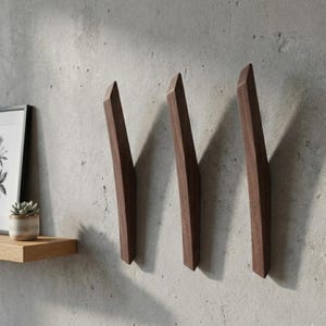 Minimalist dark walnut wooden wall hooks in a mid-century modern style, mounted vertically on a textured light gray wall, featuring three sculpted solid wood hooks with clean lines, styled with a floating wooden shelf, framed artwork, and a small potted plant