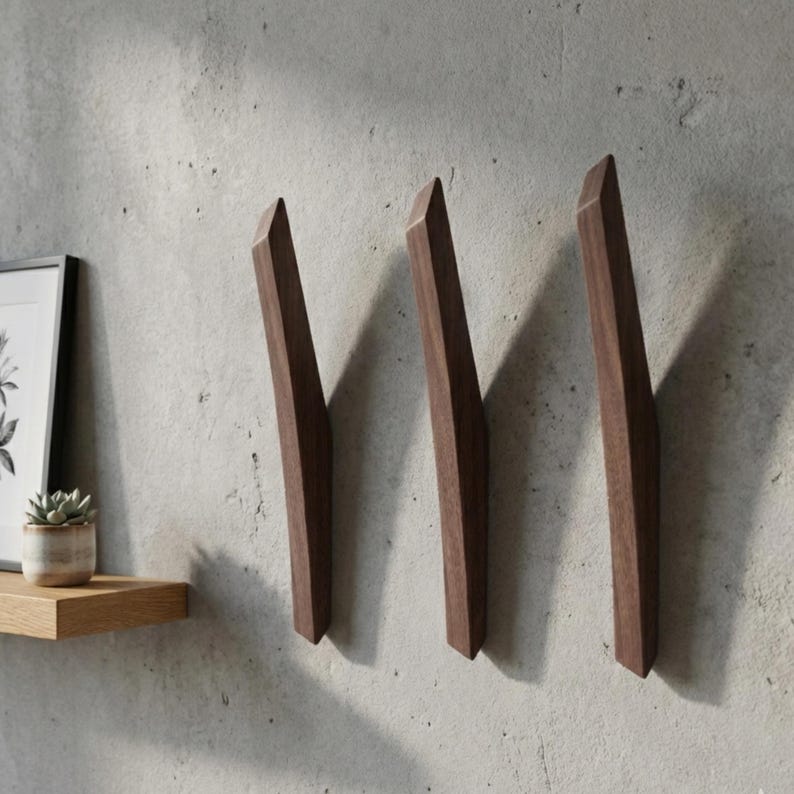 Minimalist dark walnut wooden wall hooks in a mid-century modern style, mounted vertically on a textured light gray wall, featuring three sculpted solid wood hooks with clean lines, styled with a floating wooden shelf, framed artwork, and a small potted plant
