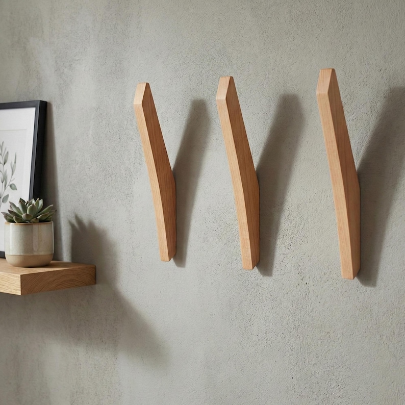 Minimalist mid-century modern wooden wall hooks mounted on a textured light gray wall, featuring three angled solid wood hooks with a natural finish, styled in a contemporary home interior with a small wooden shelf, framed art, and a potted plant