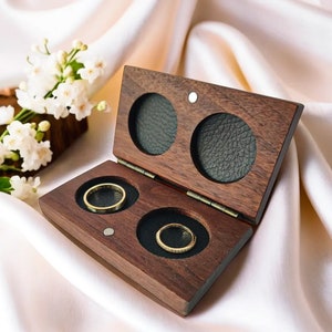 May include: A wooden ring box with two compartments, each lined with black velvet. The box is open and two gold rings are visible inside.