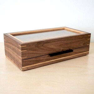 May include: A rectangular wooden storage box crafted from dark walnut wood with light wood accents at the corners and edges. The box features a clear glass lid on top and a single drawer below with a thin, horizontal black metal handle.