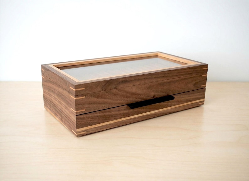 May include: A rectangular wooden storage box crafted from dark walnut wood with light wood accents at the corners and edges. The box features a clear glass lid on top and a single drawer below with a thin, horizontal black metal handle.