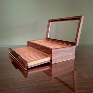May include: A dark brown wooden display box with a hinged glass lid and a pull-out drawer. The interior features light-colored wood slats arranged in parallel rows for storage. The box has a rectangular shape with visible joinery at the corners.