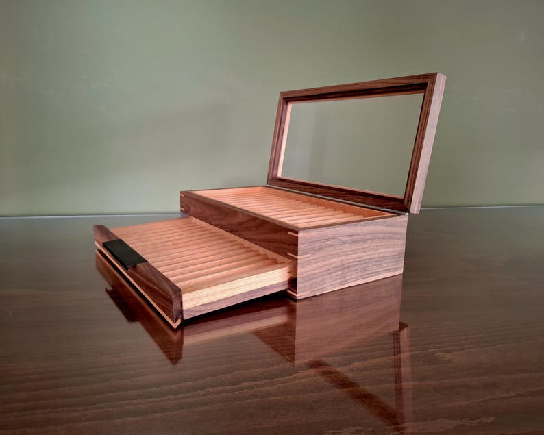 May include: A dark brown wooden display box with a hinged glass lid and a pull-out drawer. The interior features light-colored wood slats arranged in parallel rows for storage. The box has a rectangular shape with visible joinery at the corners.