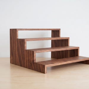 May include: A wooden display stand with three stepped shelves. The stand is made of dark brown wood and has a modern, minimalist design. The shelves are open, providing space for displaying items. The stand is suitable for showcasing products.