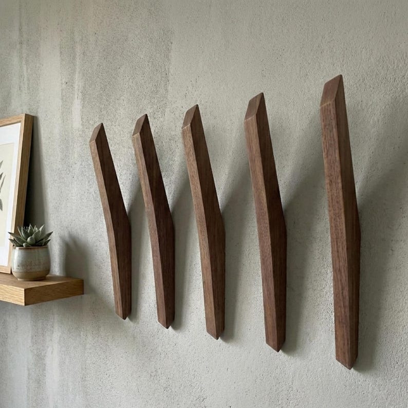Mid-century modern dark walnut wooden wall hooks arranged in a row on a textured light gray wall, featuring five sculpted solid wood hooks with clean lines, styled with a floating wooden shelf, framed artwork, and a small potted plant for a minimalist home interior