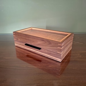 May include: A rectangular wooden box featuring a dark brown walnut grain, light-colored wood inlay accents, and visible finger joints at the corners. The lid includes a clear glass panel, and a thin black horizontal handle is centered on the front.