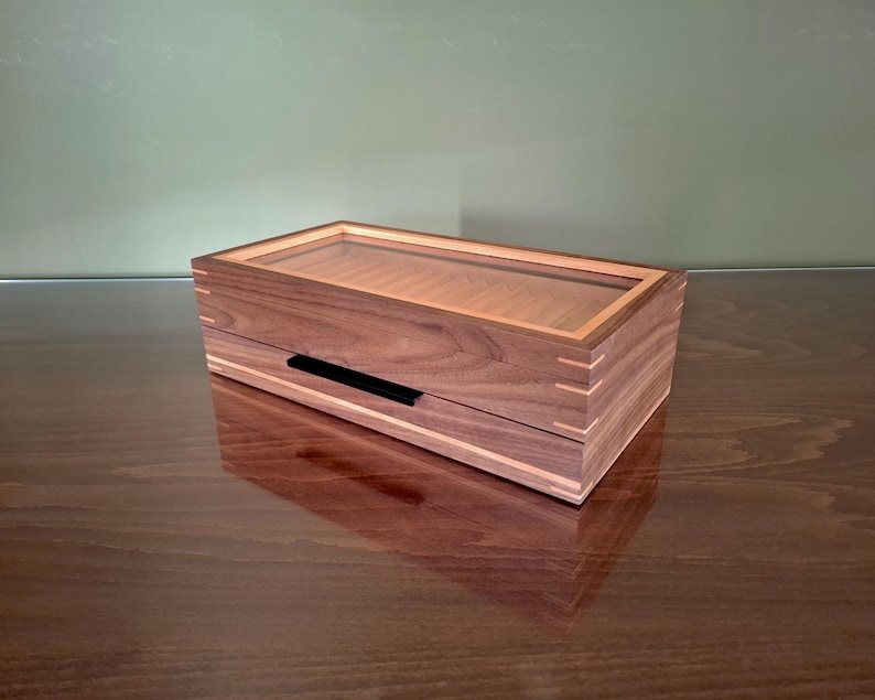 May include: A rectangular wooden box featuring a dark brown walnut grain, light-colored wood inlay accents, and visible finger joints at the corners. The lid includes a clear glass panel, and a thin black horizontal handle is centered on the front.