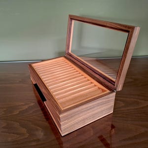 May include: A rectangular wooden display box with a hinged lid featuring a clear glass panel. The interior contains a series of parallel, light-colored wooden grooves designed for organizing items, while the exterior displays a dark wood grain.