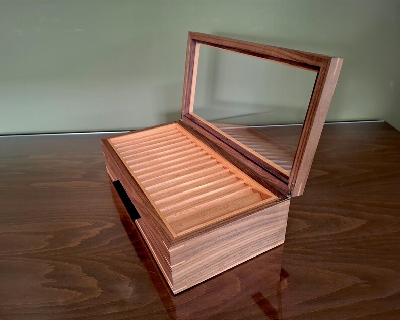 May include: A rectangular wooden display box with a hinged lid featuring a clear glass panel. The interior contains a series of parallel, light-colored wooden grooves designed for organizing items, while the exterior displays a dark wood grain.