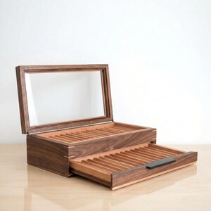 May include: A dark brown wooden display box with a hinged glass lid and a pull-out drawer. The interior features multiple grooved slots for organizing items, with a small black handle on the front of the drawer and visible wood grain.