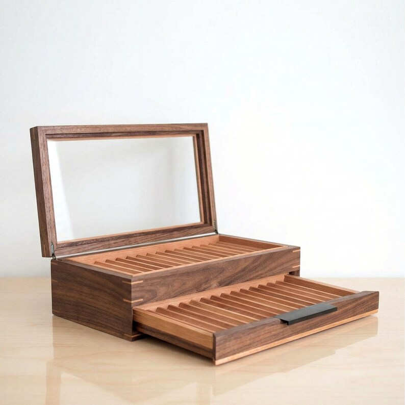 May include: A dark brown wooden display box with a hinged glass lid and a pull-out drawer. The interior features multiple grooved slots for organizing items, with a small black handle on the front of the drawer and visible wood grain.