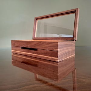 May include: A rectangular wooden box with a dark brown wood grain finish and light wood corner accents. The box features a hinged lid with a clear glass panel and a thin black horizontal handle on the front face of the lower storage compartment.
