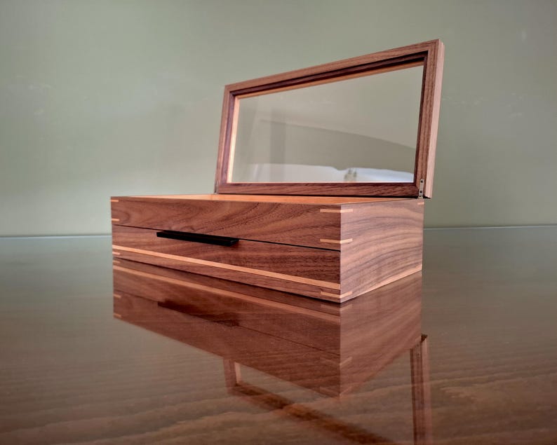 May include: A rectangular wooden box with a dark brown wood grain finish and light wood corner accents. The box features a hinged lid with a clear glass panel and a thin black horizontal handle on the front face of the lower storage compartment.