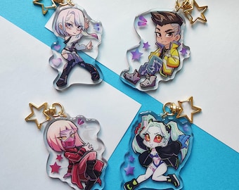 May include: Four colorful character keychains with a star charm attached to each. The characters are a girl with pink hair, a girl with blue hair, a boy with blue hair, and a girl with purple hair. The keychains are made of clear acrylic and have a glossy finish.