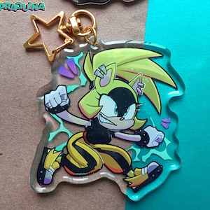 Op de afbeelding: Een geel en zwart cartoonfiguur met stekelig geel haar en een zwart masker. Het figuur staat in een vechtpose met een geel en zwart pak. Het figuur zit op een transparante acryl sleutelhanger met een gouden sterbedel.